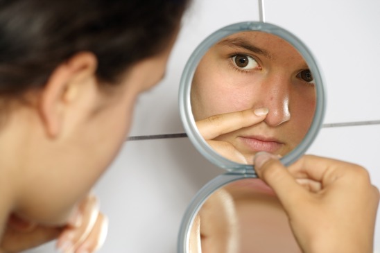 Young teenage female holding a mirror looking at her pimple with concern.