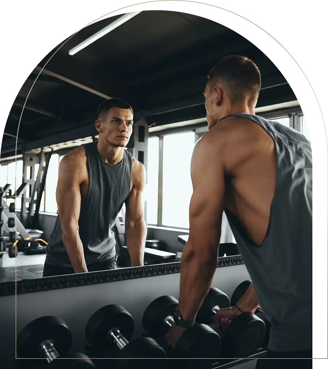 Muscular man in sleeveless shirt looking at reflection in gym mirror Man in gym looking at mirror with dumbbells